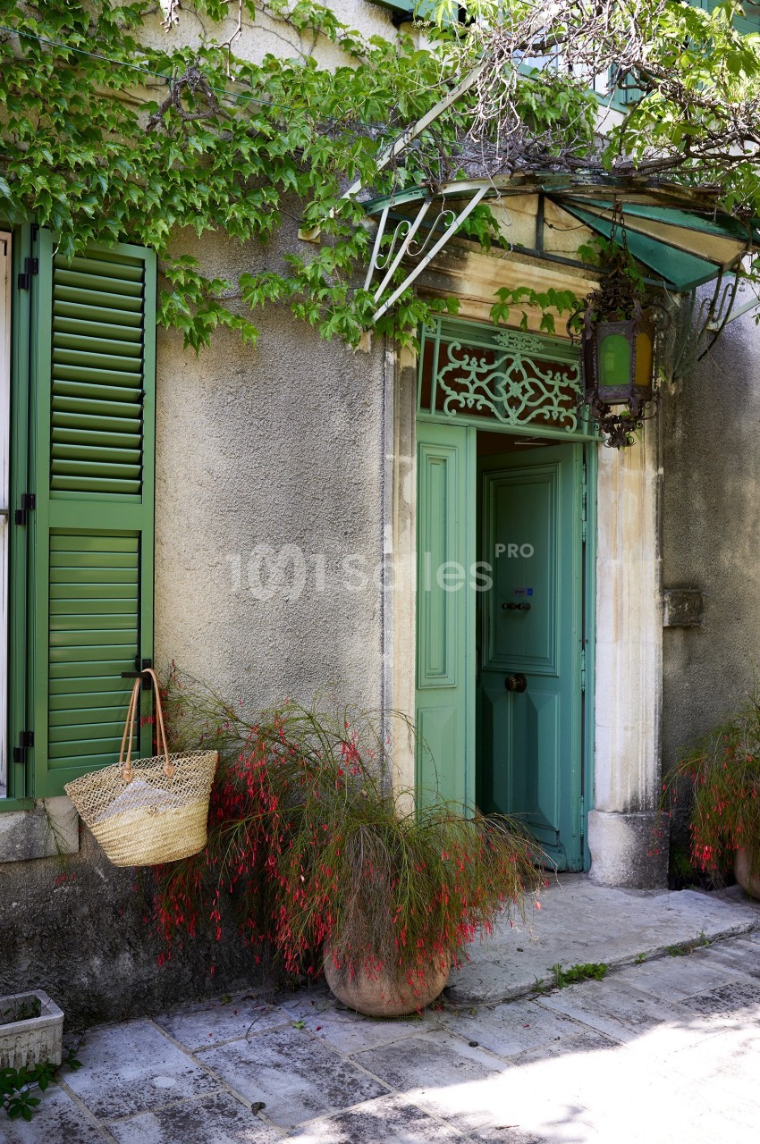 Entrée de maison avec porte et volets verts, plantes en pots et panier suspendu sous une marquise en métal.