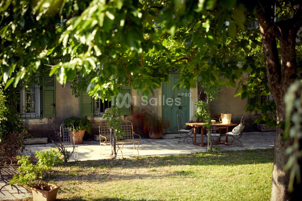 Terrasse ombragée avec mobilier de jardin en métal, devant une maison aux volets verts entourée de végétation.