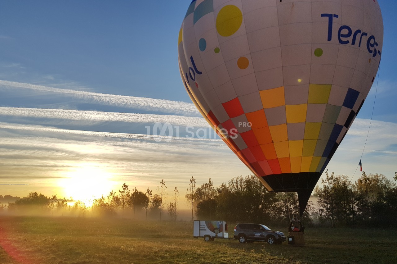Montgolfière colorée en cours de gonflage dans un champ au lever du soleil, avec une voiture et une remorque à proximité.