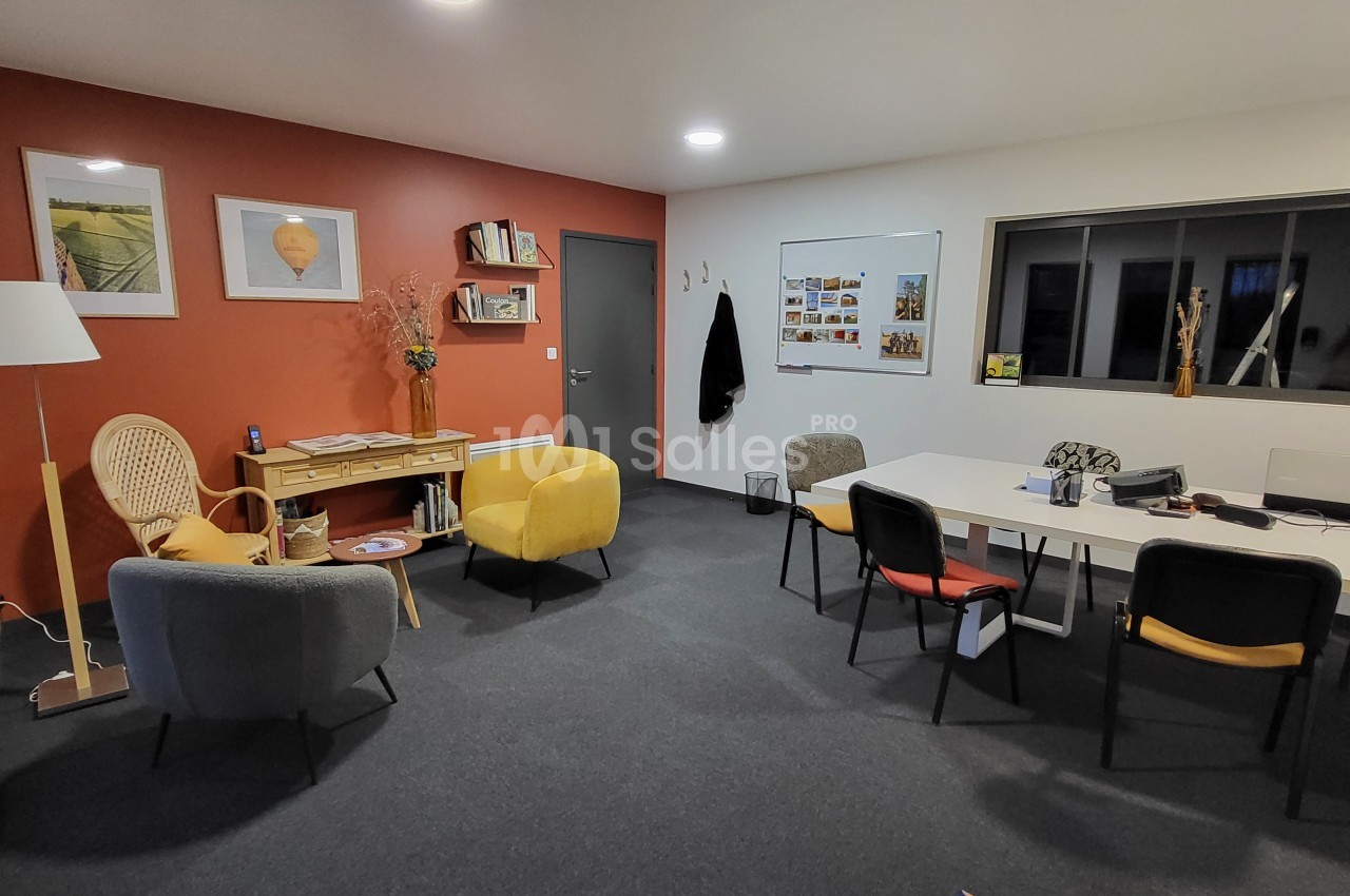 Salle de réunion lumineuse avec fauteuils, table blanche, étagères, mur rouge et tableau d'affichage.