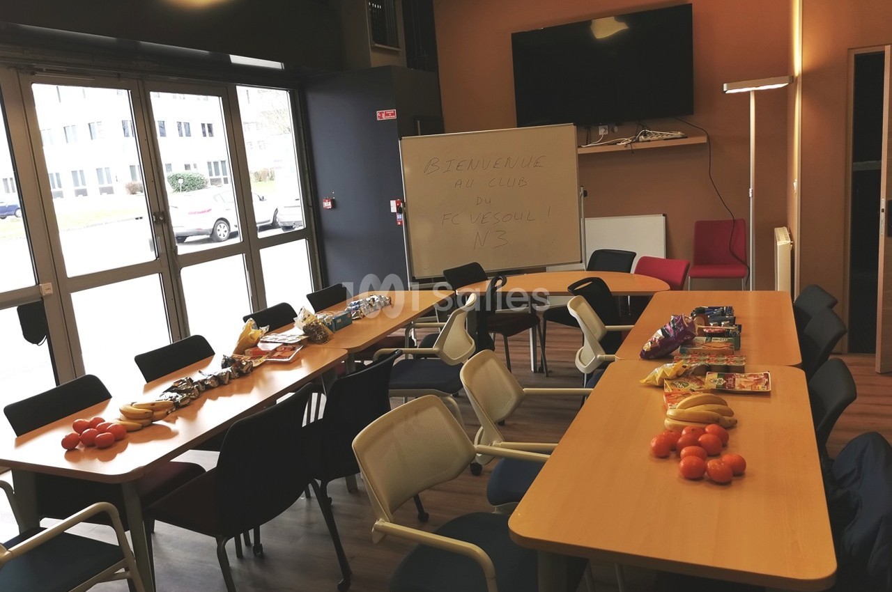 Salle de réunion avec tables disposées en U, garnies de fruits et collations, tableau blanc et chaises autour.