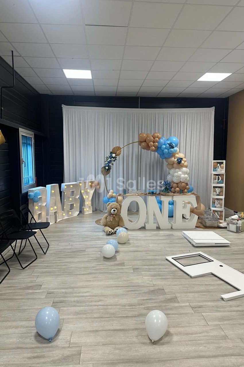 Décoration de fête avec ballons bleus, blancs et beiges, lettres géantes ’BABY ONE’ et un ours en peluche.