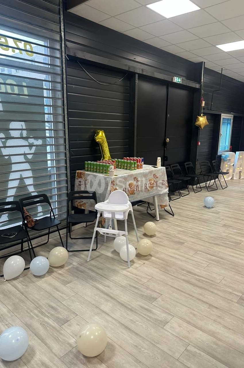 Salle décorée pour une fête avec ballons, table ornée de nappes à motif ’baby’ et chaises alignées.