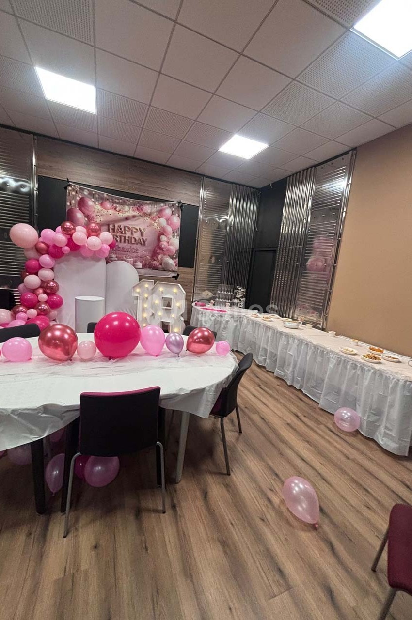 Salle décorée pour un anniversaire avec ballons roses, table dressée et panneau ’Happy Birthday’ en arrière-plan.