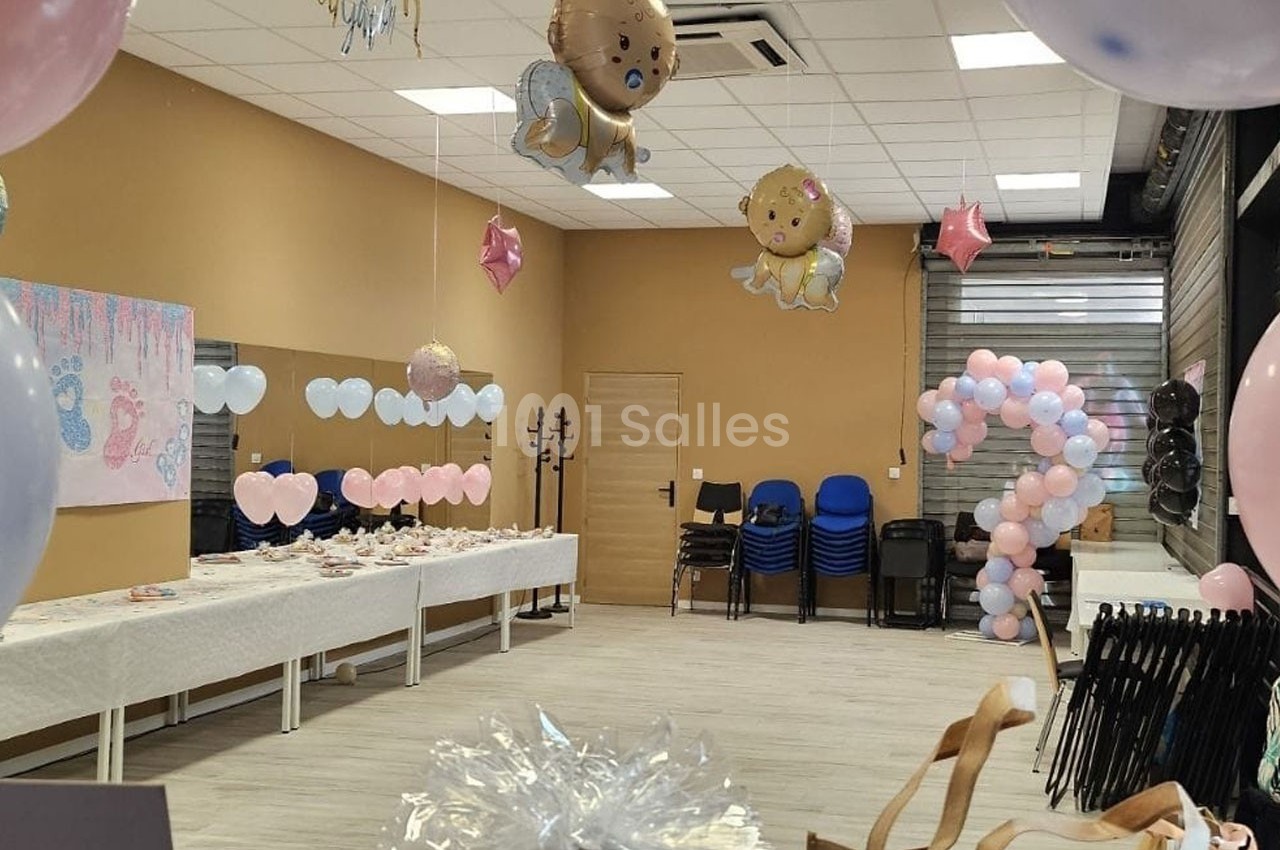 Salle décorée pour une fête prénatale avec ballons, table dressée et chaises empilées au fond.