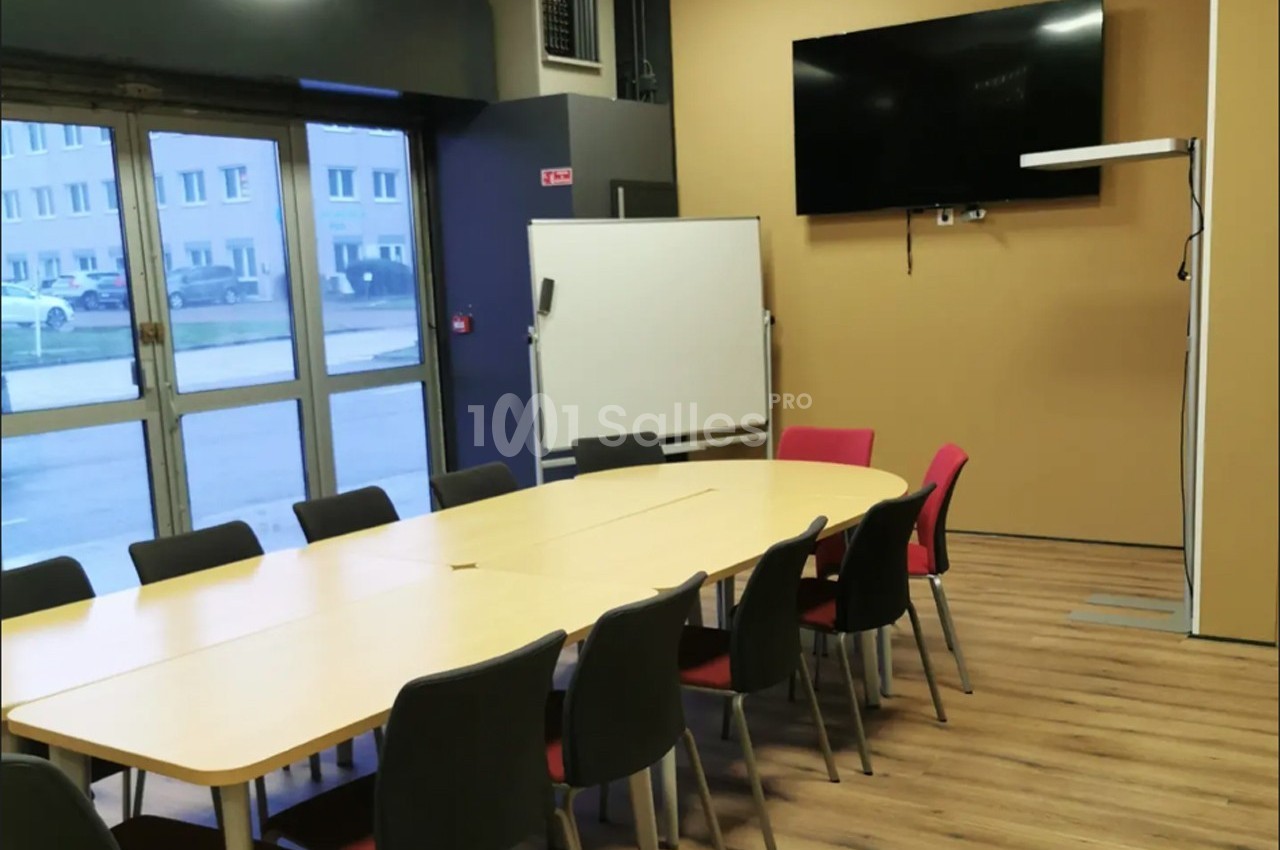 Salle de réunion avec une grande table, des chaises, un tableau blanc, un écran mural et un sol en parquet.