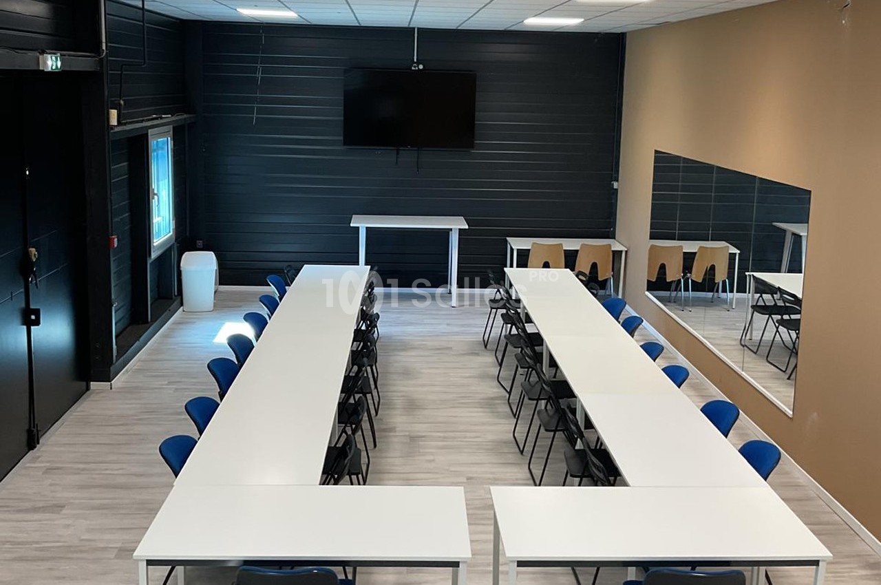 Salle de réunion avec tables disposées en U, chaises bleues, écran mural, miroir et sol en parquet clair.