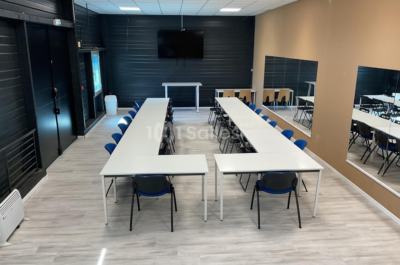 Salle de réunion avec tables disposées en U, chaises bleues, écran mural et murs partiellement recouverts de miroirs.