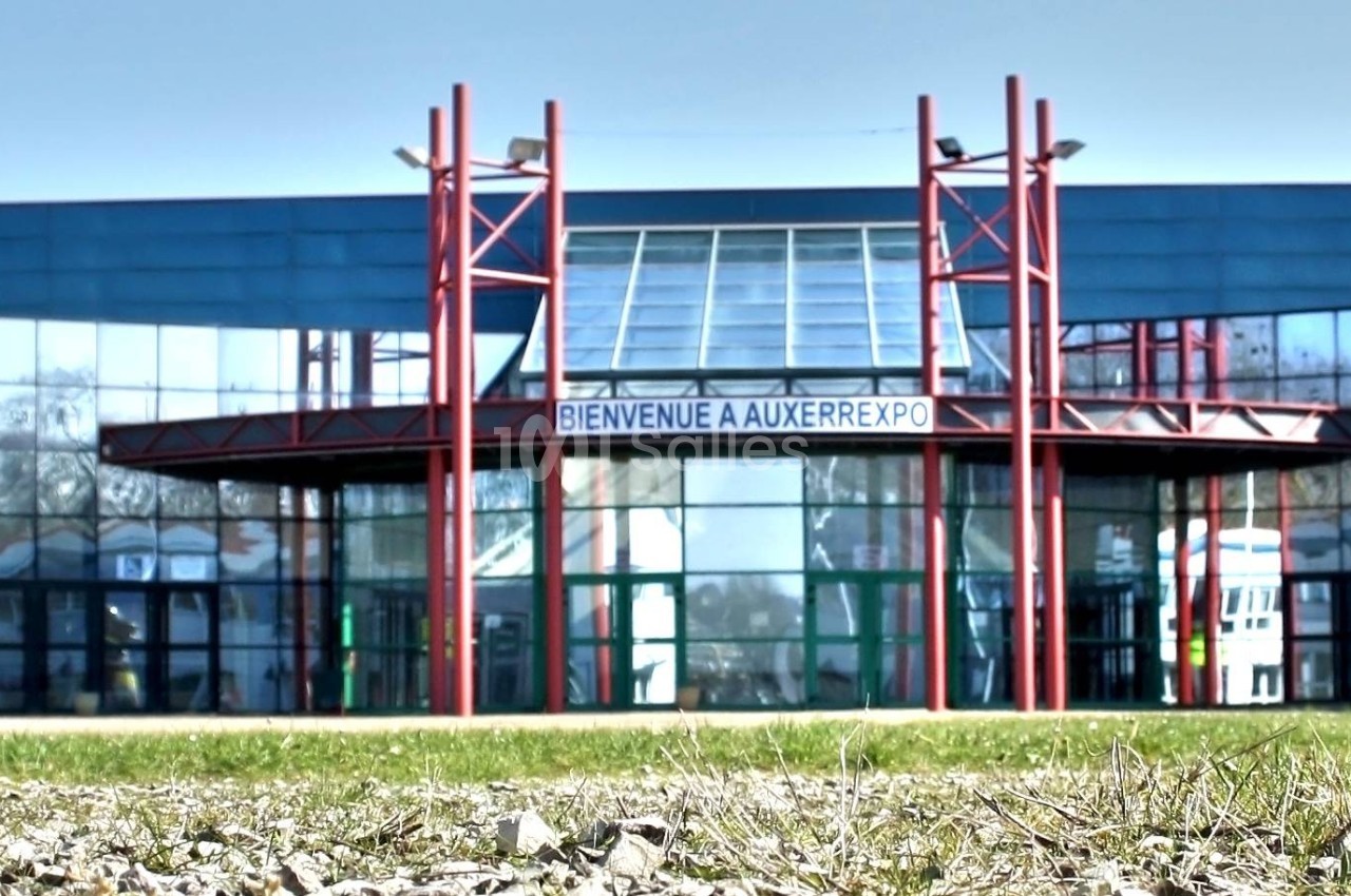 Façade vitrée d'un bâtiment moderne avec une entrée centrale et des structures rouges, vue depuis un espace extérieur.