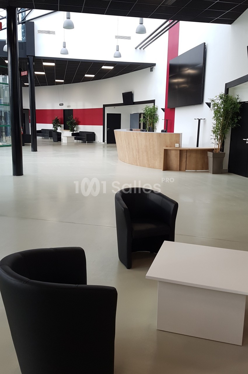 Espace d'accueil moderne avec fauteuils noirs, table basse blanche, comptoir en bois et écrans muraux.