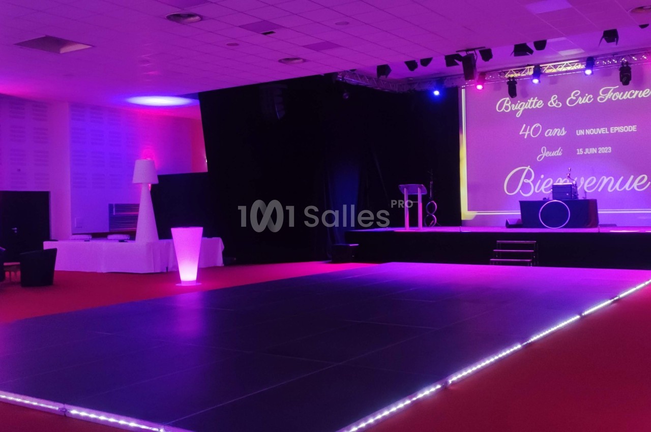 Salle de réception avec piste de danse éclairée, scène équipée et écran affichant un message de bienvenue.