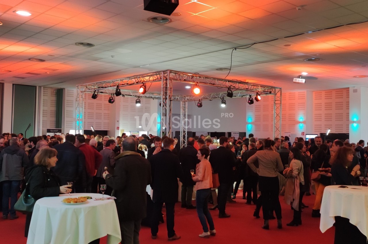 Salle de réception avec de nombreuses personnes debout discutant sous un éclairage coloré, tables hautes en premier plan.