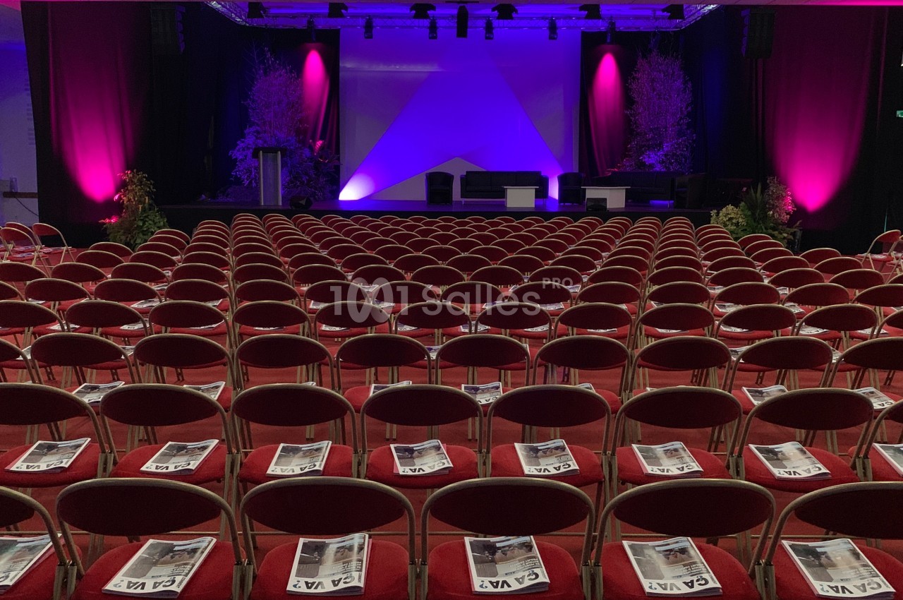Salle de conférence avec des rangées de chaises rouges, des brochures posées sur chaque siège et une scène éclairée en…
