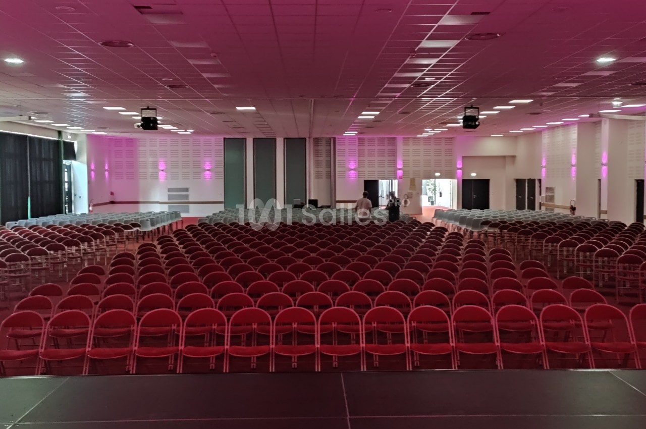 Salle de conférence avec rangées de chaises rouges alignées, éclairage rose et scène au premier plan.