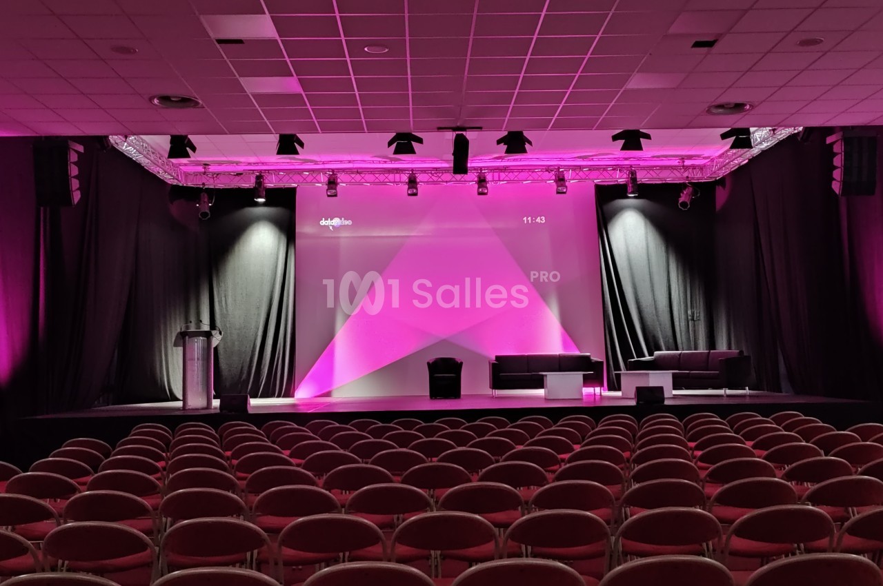 Salle de conférence avec scène éclairée en rose, podium, fauteuils et rangées de chaises vides au premier plan.