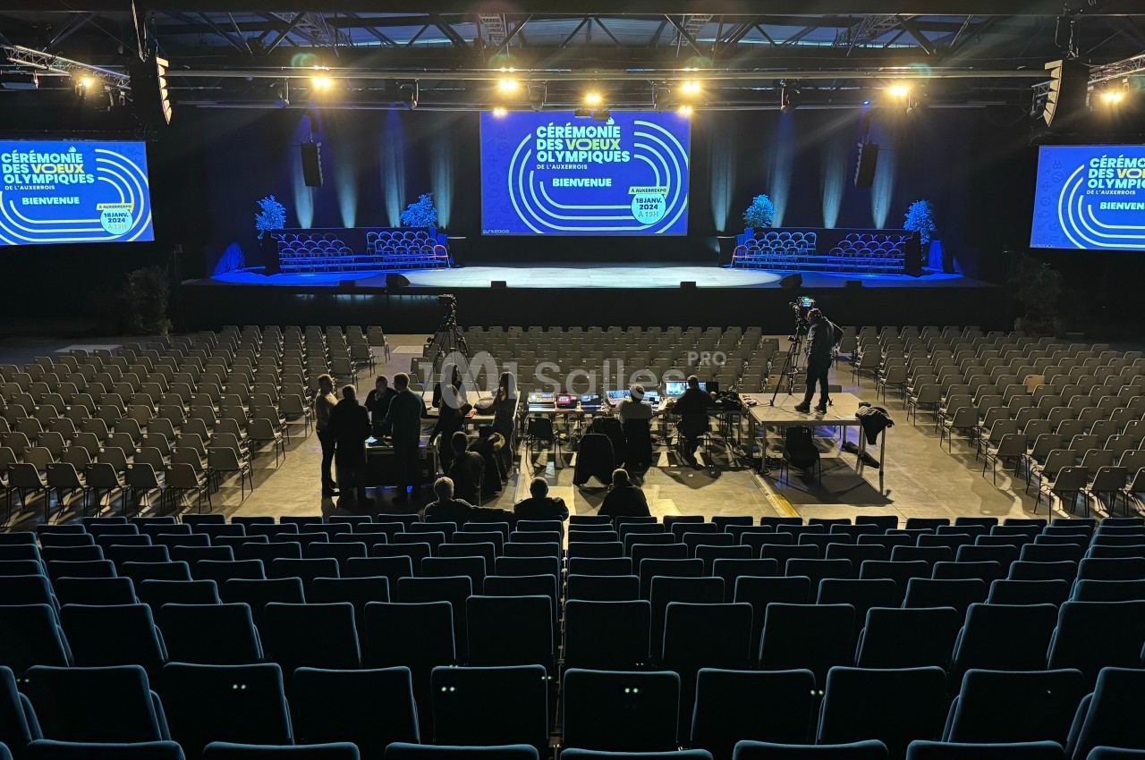 Salle de spectacle avec scène éclairée, écrans affichant ’Cérémonie des Vœux Olympiques’, et rangées de sièges vides.