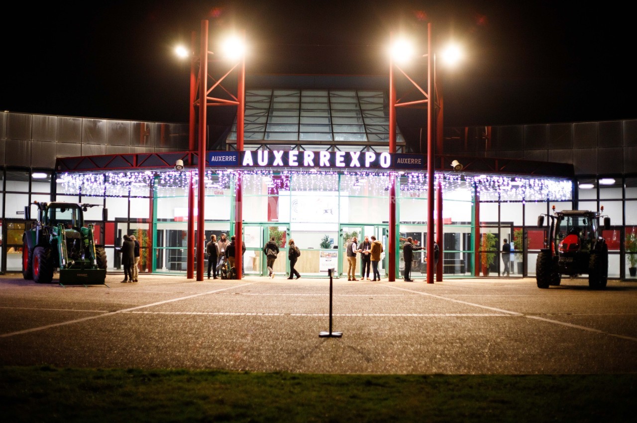 Façade illuminée du bâtiment Auxerrexpo de nuit, avec des visiteurs entrant et deux tracteurs exposés devant.