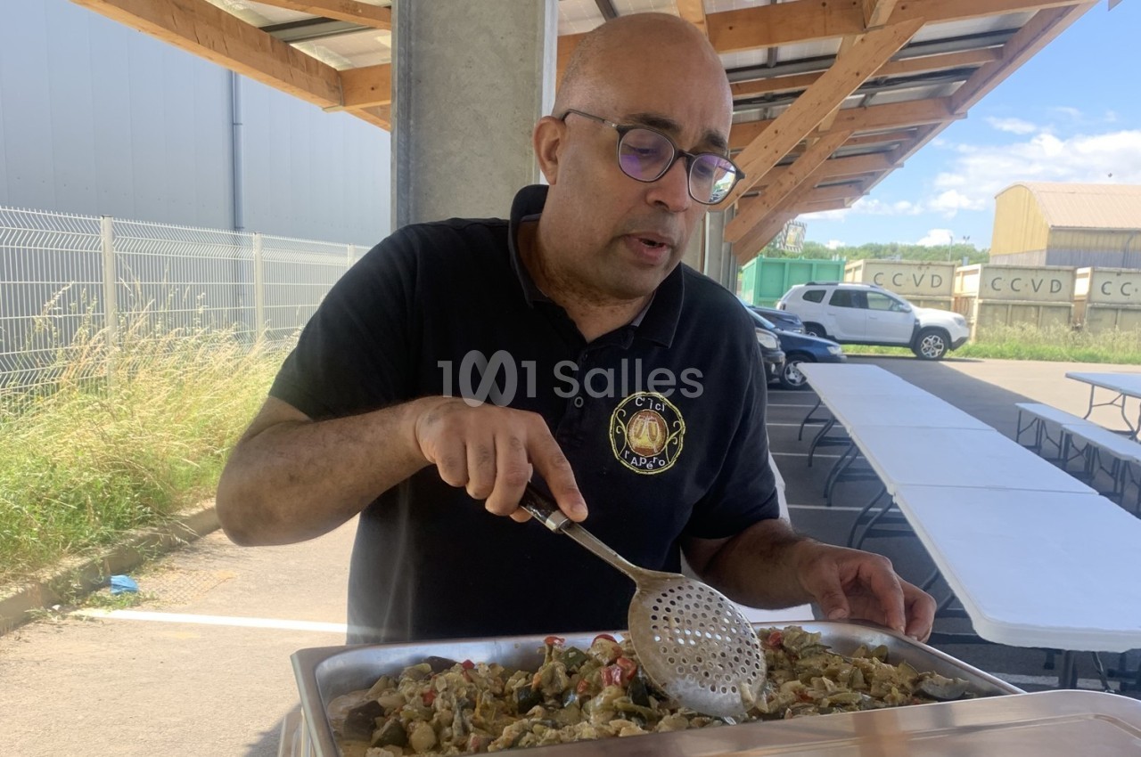 Un homme sert un plat chaud à l'aide d'une écumoire sous un abri extérieur, près de tables vides.