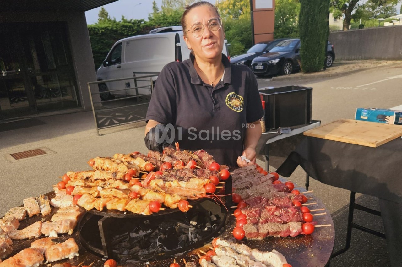 Une femme prépare des brochettes variées sur un barbecue rond en extérieur, avec des voitures en arrière-plan.