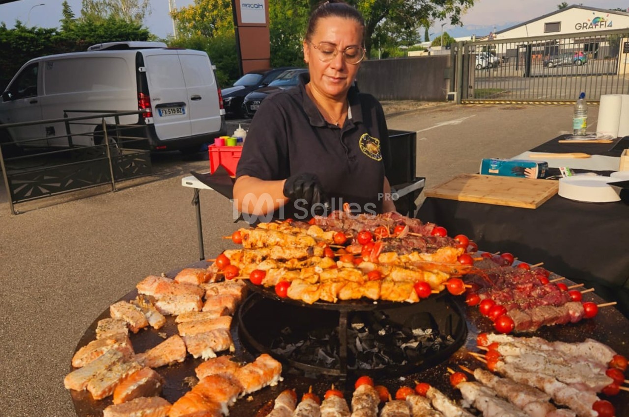 Une femme cuisine des brochettes variées sur un barbecue circulaire en plein air, entourée d'ingrédients.
