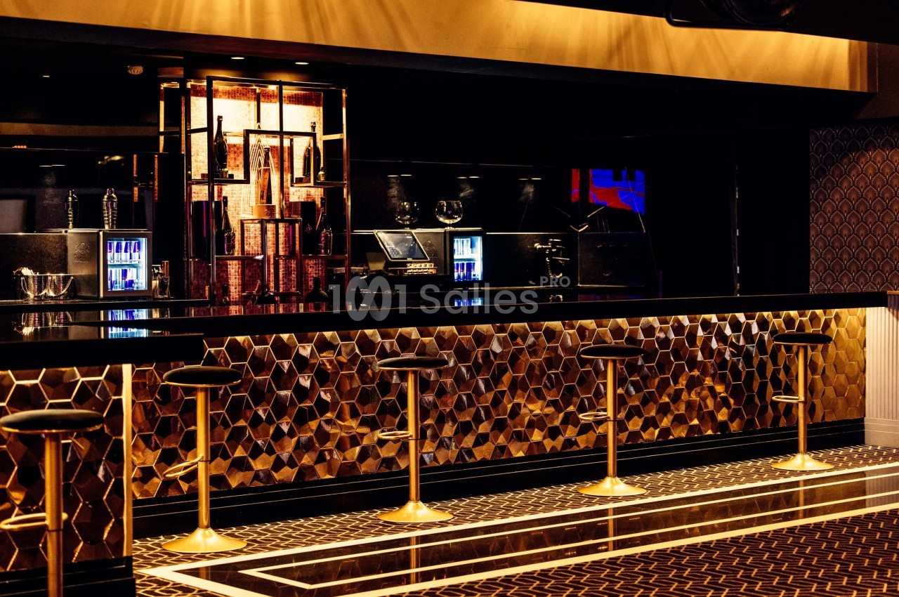 Bar moderne avec comptoir doré à motifs géométriques, tabourets alignés et éclairage tamisé.