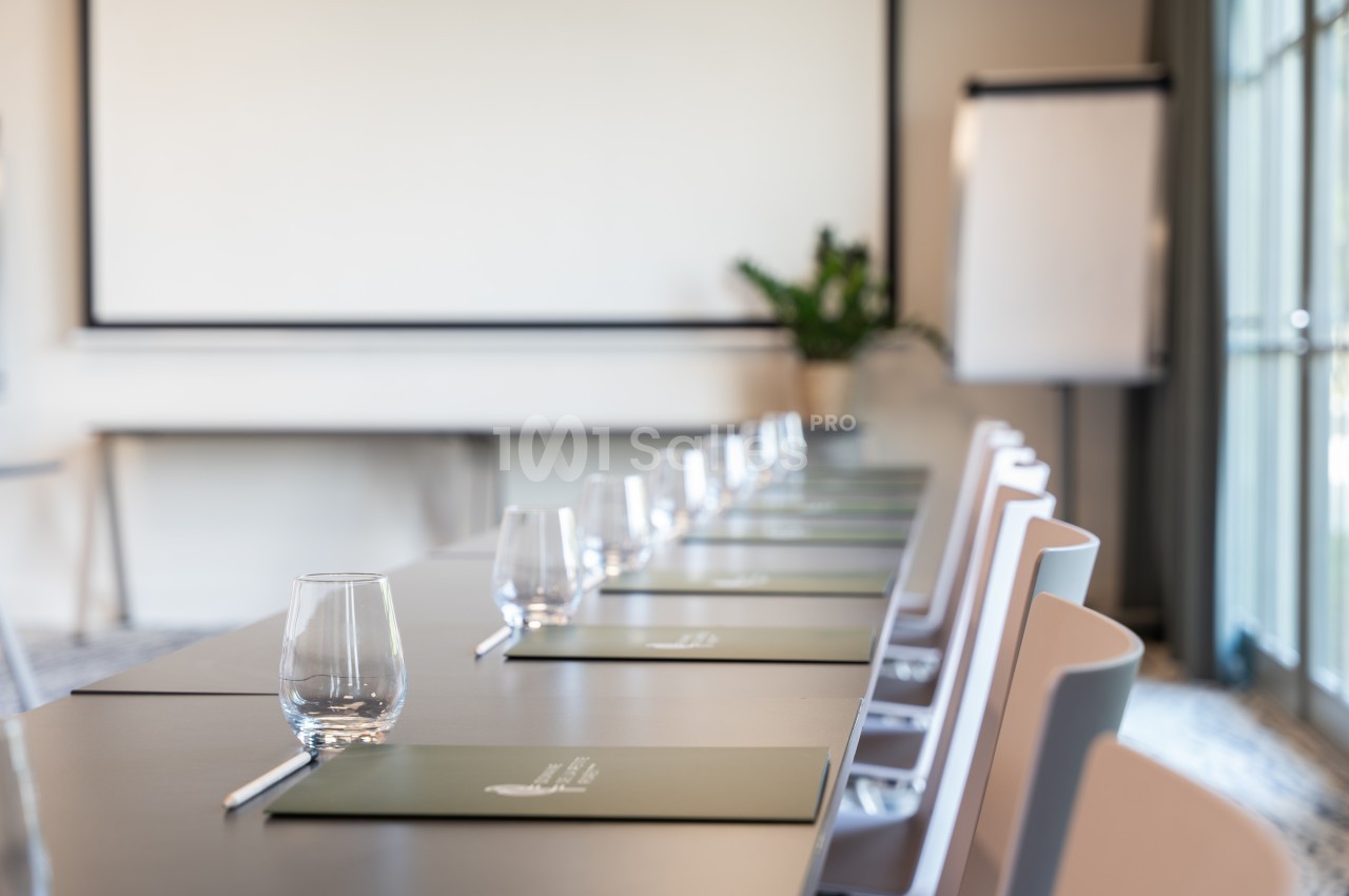 Salle de réunion lumineuse avec une table alignée de dossiers verts, verres et stylos, face à un écran et un tableau blanc.