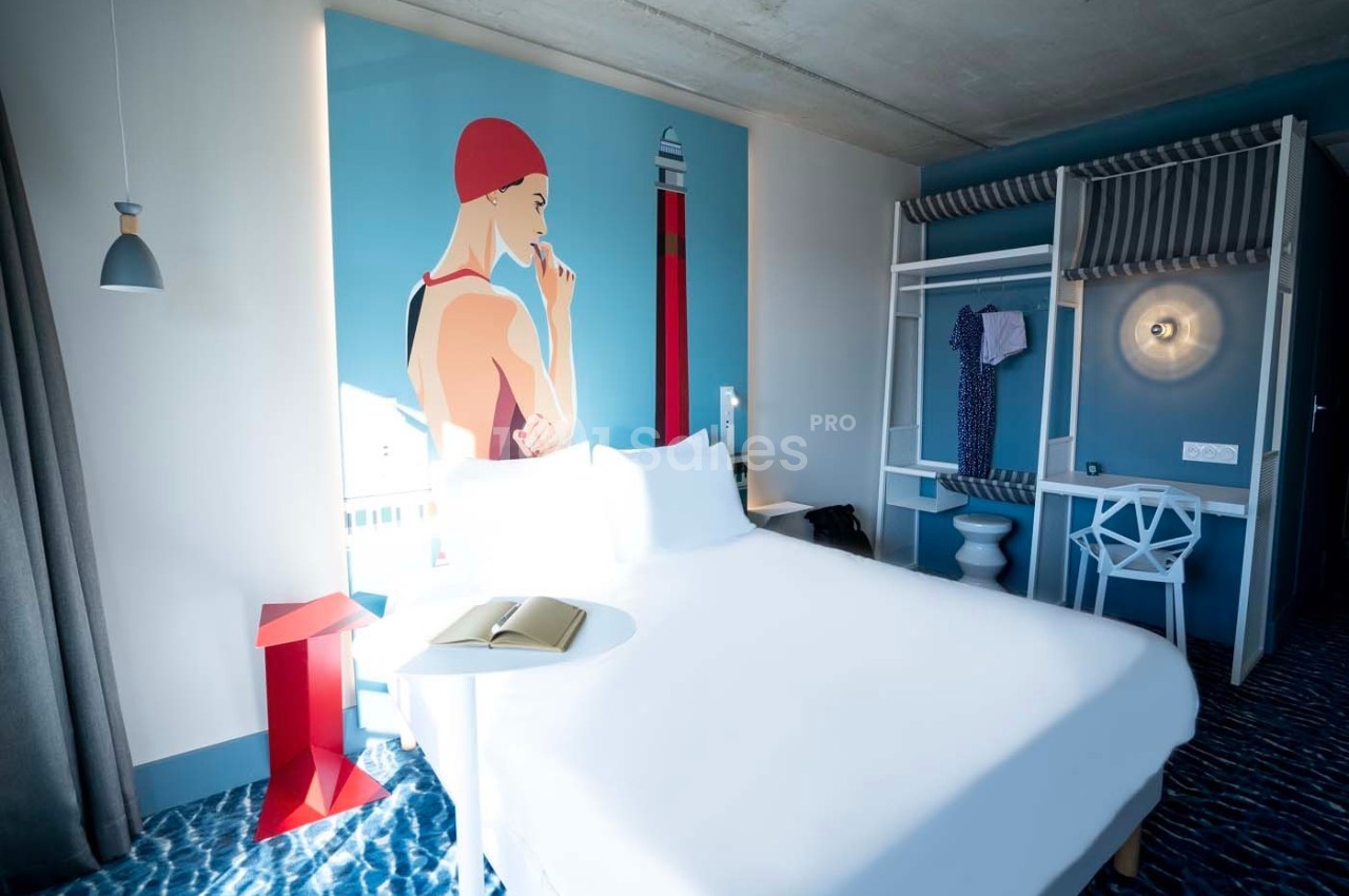Chambre d'hôtel moderne avec lit double, décoration murale colorée, penderie ouverte et lumière naturelle.