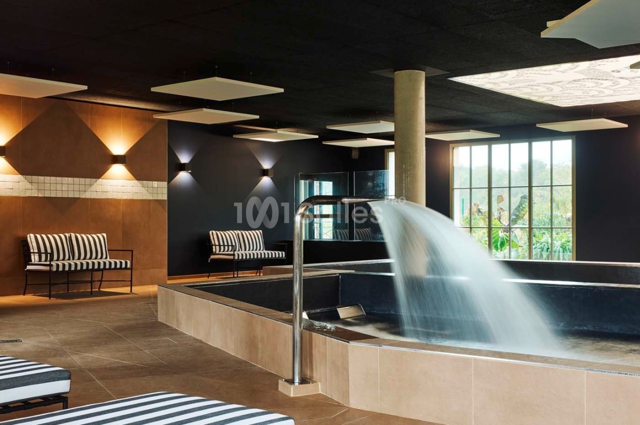 Salle de spa avec bassin, cascade d'eau, chaises longues rayées et grandes fenêtres donnant sur un jardin.