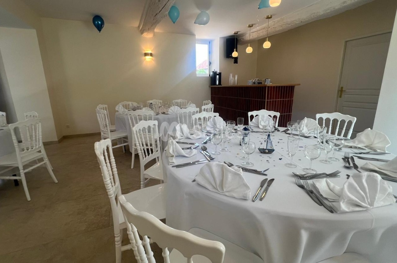 Salle de réception lumineuse avec tables rondes dressées, nappes blanches, serviettes pliées et chaises assorties.