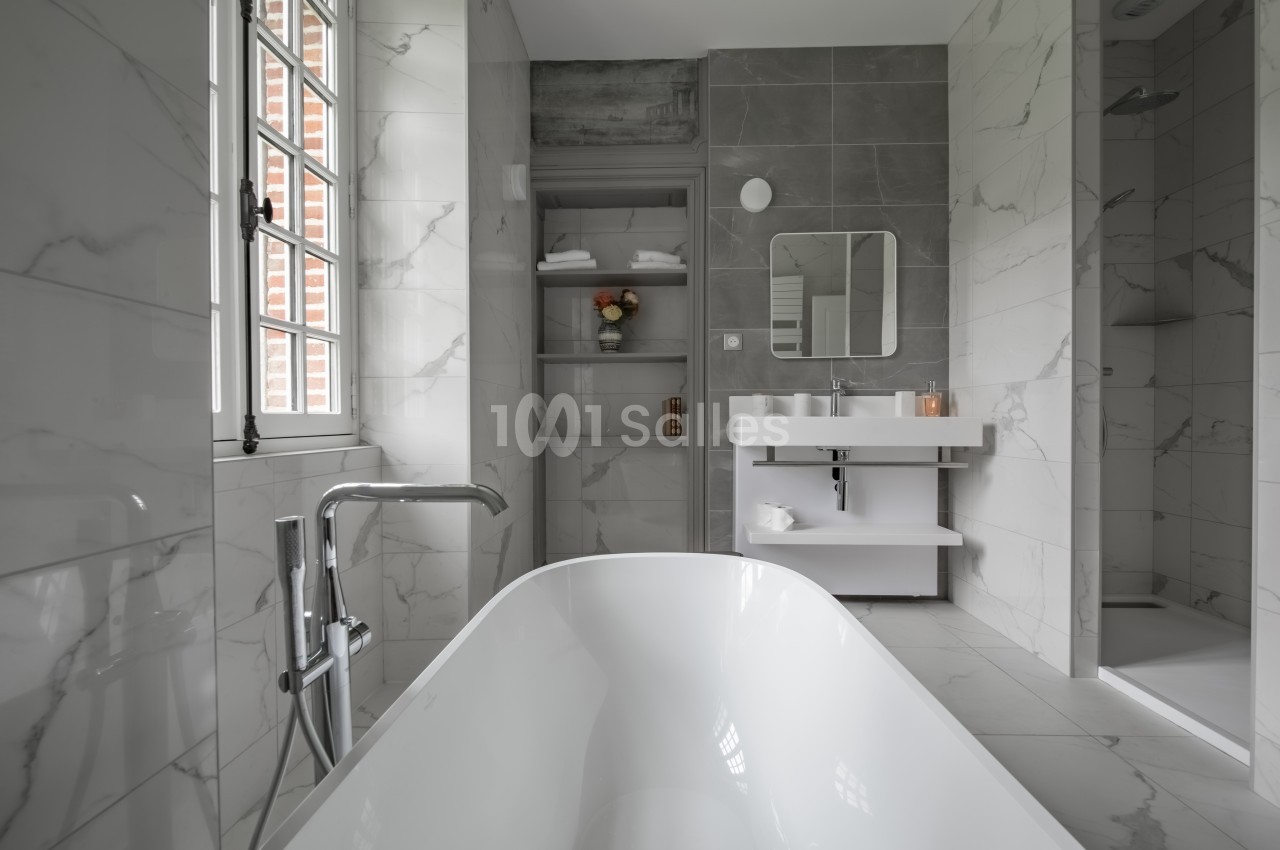 Salle de bain moderne avec baignoire, lavabo double, douche et étagères, murs en marbre et carrelage gris.