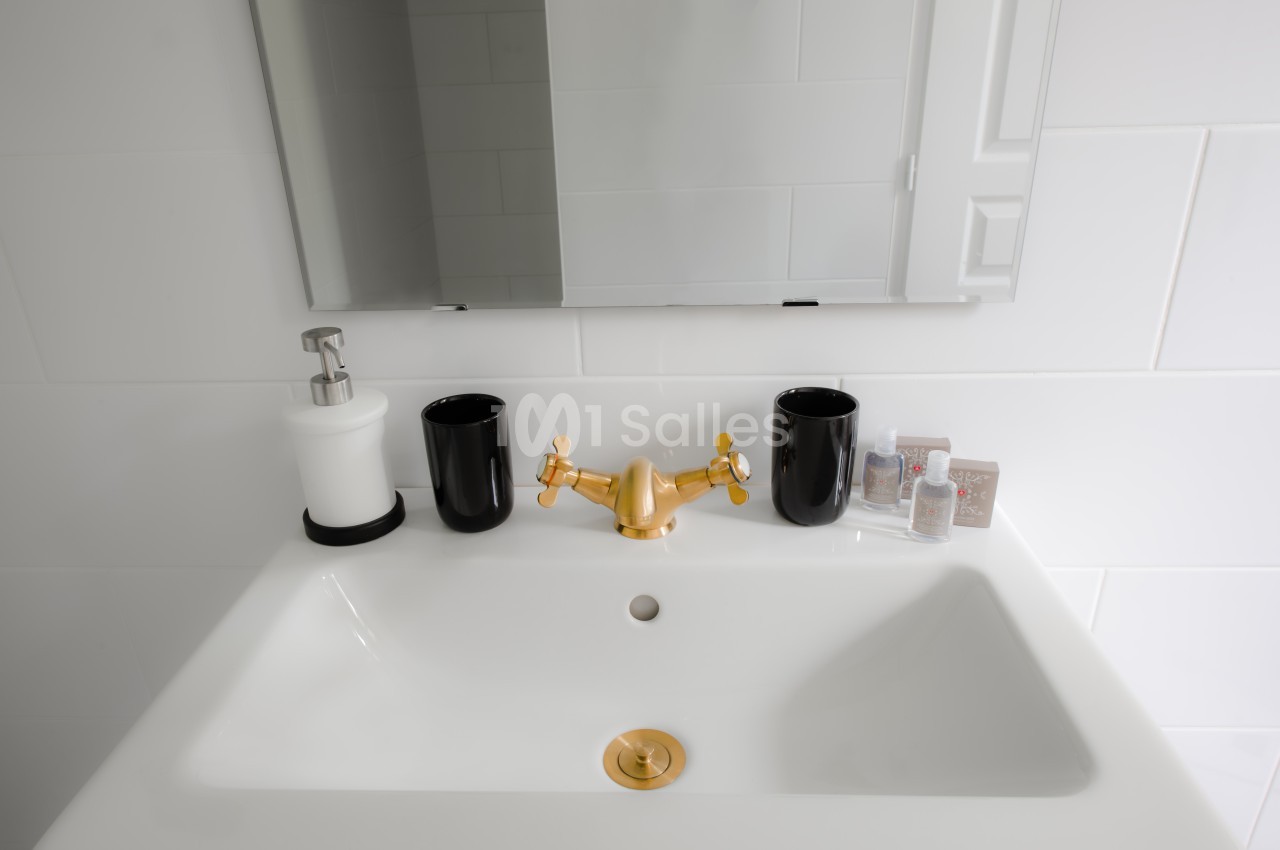 Lavabo blanc avec robinet doré, deux gobelets noirs, un distributeur de savon et un miroir mural.