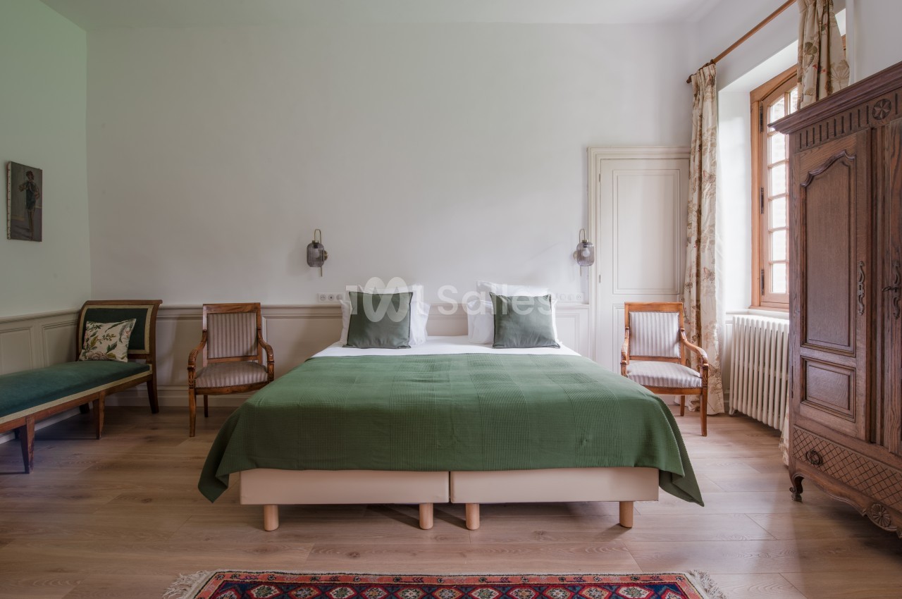 Chambre lumineuse avec lit double, couvre-lit vert, fauteuils en bois, armoire ancienne et tapis coloré.