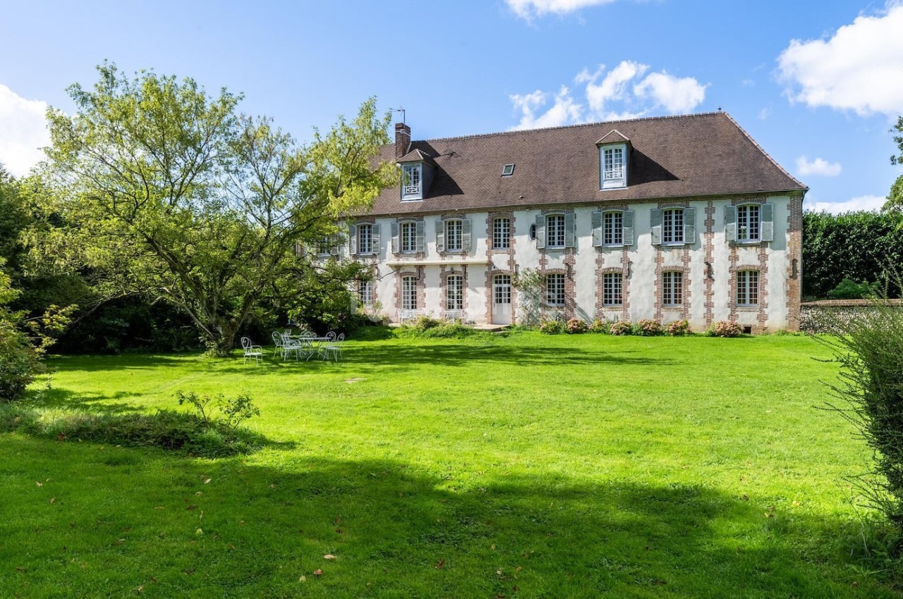 Manoir en pierre avec toit en ardoise, entouré d'un grand jardin verdoyant sous un ciel bleu.