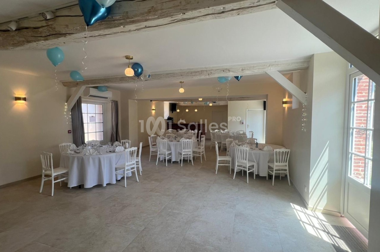 Salle de réception lumineuse décorée de ballons bleus, avec tables rondes dressées et chaises blanches.