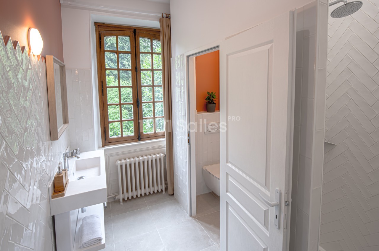 Salle de bain lumineuse avec lavabo, douche, toilettes séparées et fenêtre donnant sur un espace verdoyant.