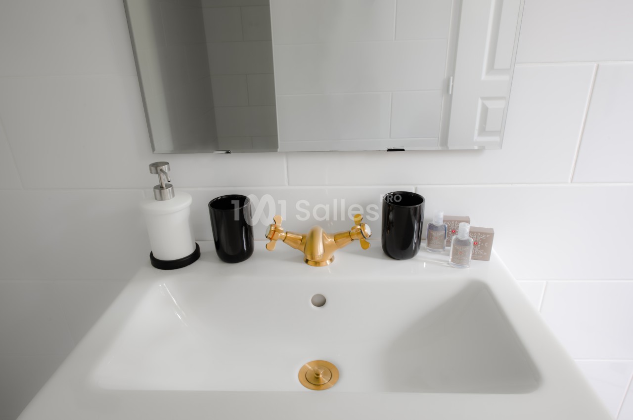 Lavabo blanc avec robinet doré, deux gobelets noirs, un distributeur de savon et un miroir mural.