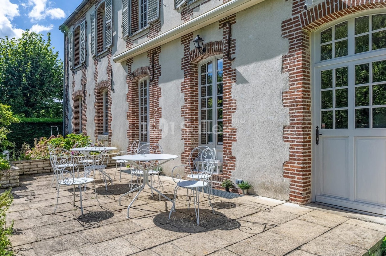 Terrasse en pierre avec tables et chaises en fer forgé devant une maison en briques et crépi, entourée de verdure.