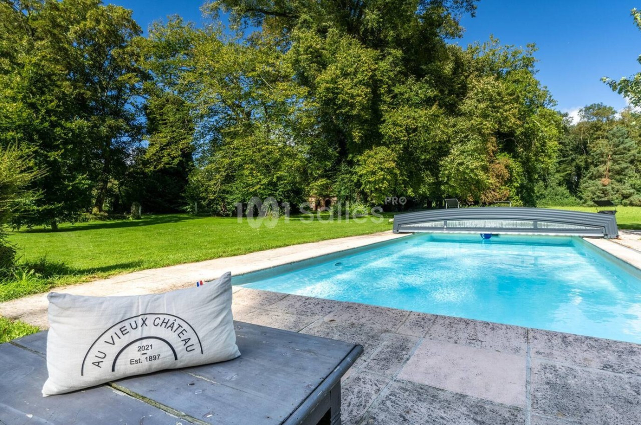 Piscine extérieure entourée de verdure avec un coussin posé sur une table en pierre au premier plan.