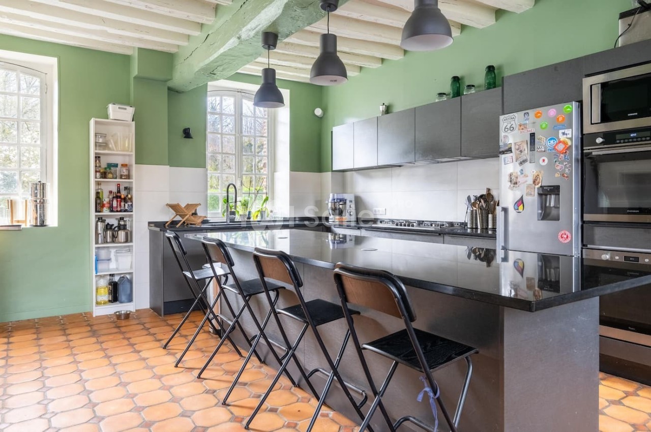 Cuisine lumineuse avec murs verts, îlot central noir, tabourets pliants, électroménagers intégrés et sol en carreaux orange.