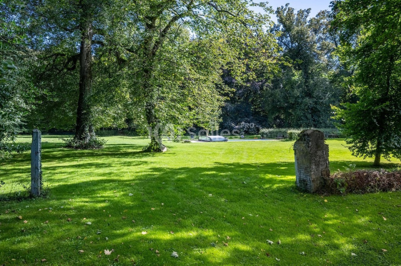 Pelouse verdoyante entourée d'arbres, avec une pierre tombale et un monument en arrière-plan sous un ciel dégagé.