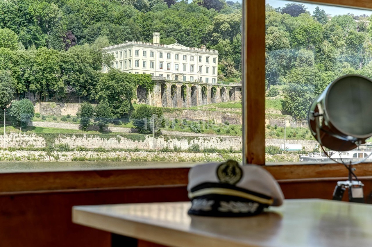 Vue d'un manoir entouré de verdure depuis l'intérieur d'un bateau, avec une casquette de marin au premier plan.