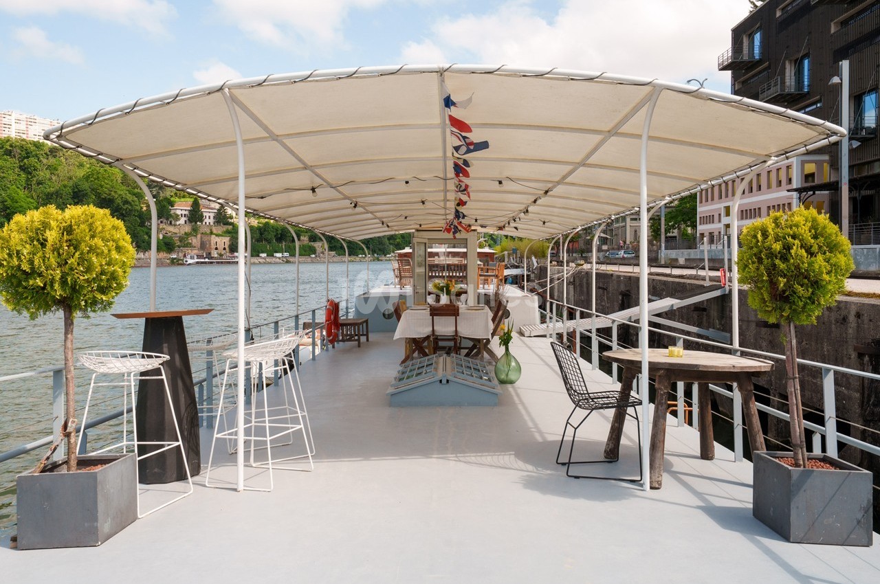 Terrasse aménagée d'un bateau avec tables, chaises et plantes, amarré près d'une rive bordée de bâtiments.