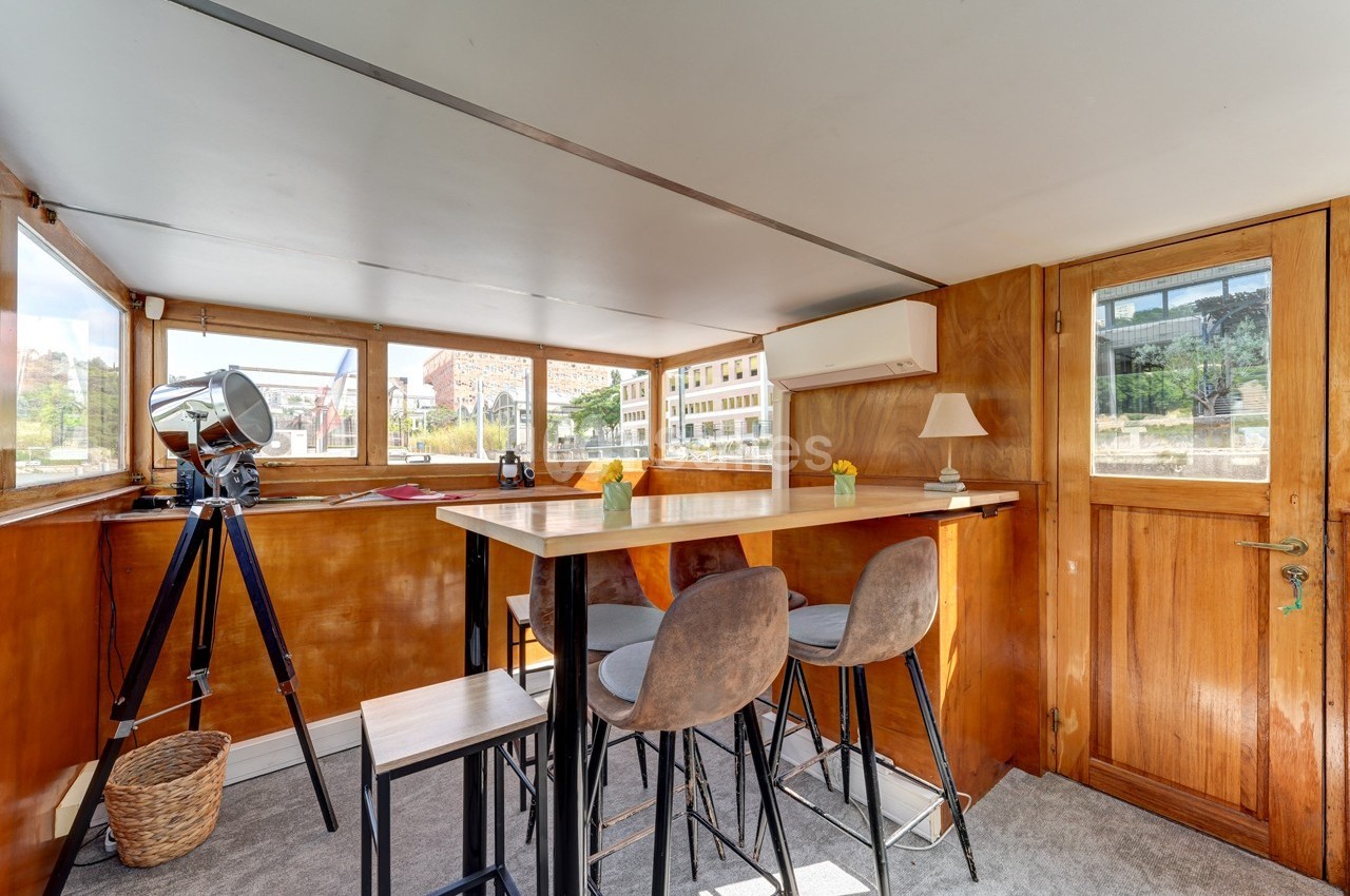 Intérieur lumineux d'une péniche avec table haute, tabourets, télescope et vue sur des bâtiments à travers les fenêtres.