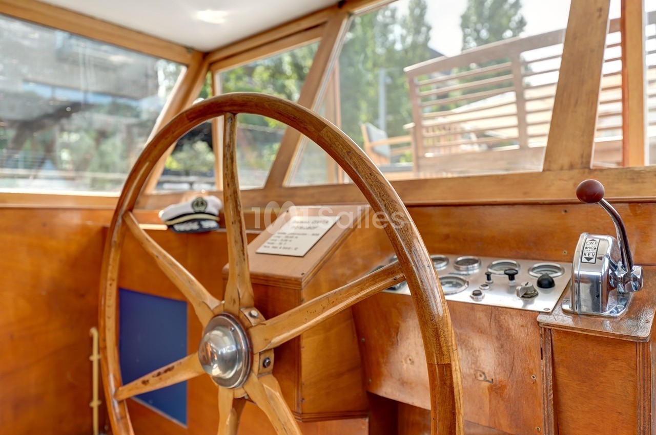 Poste de pilotage en bois d'un bateau avec un grand volant, des commandes et une vue sur l'extérieur.