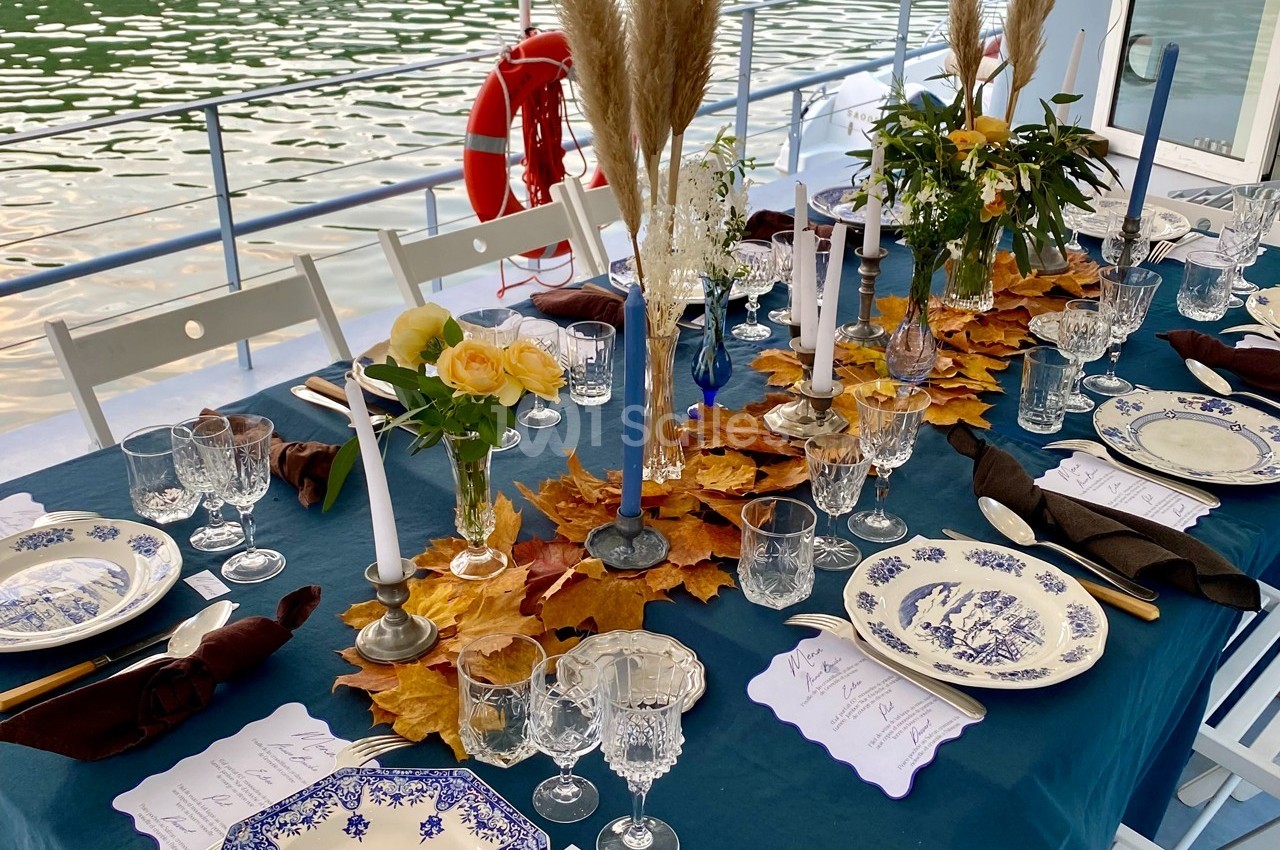 Table élégamment dressée sur une péniche, ornée de feuilles d'automne, bougies et vaisselle en porcelaine bleue.
