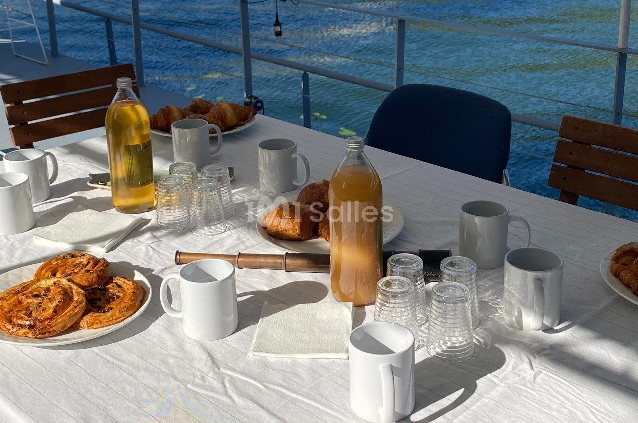 Table dressée en extérieur avec viennoiseries, quiches, bouteilles de jus, tasses et verres, près d'un plan d'eau.