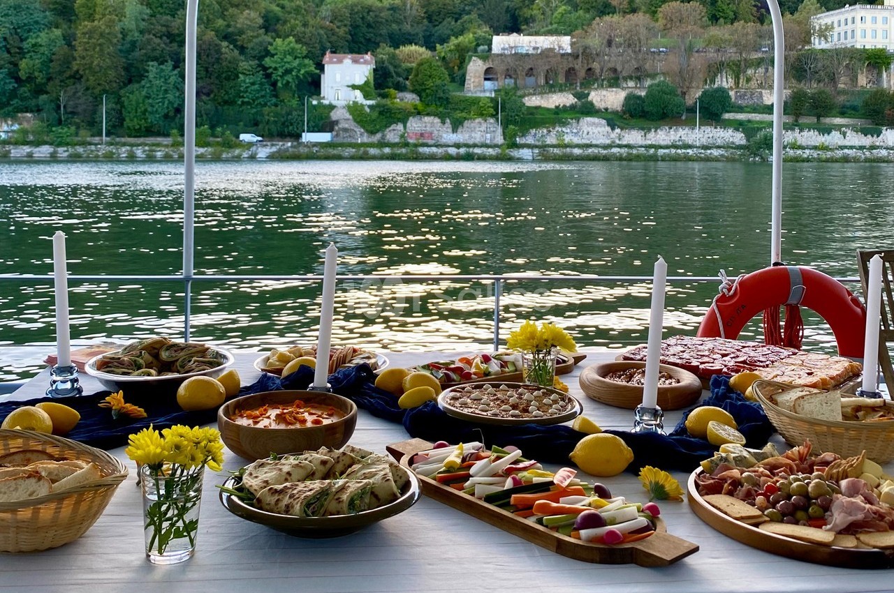 Table dressée avec divers plats et apéritifs, vue sur une rivière et des bâtiments entourés de verdure en arrière-plan.