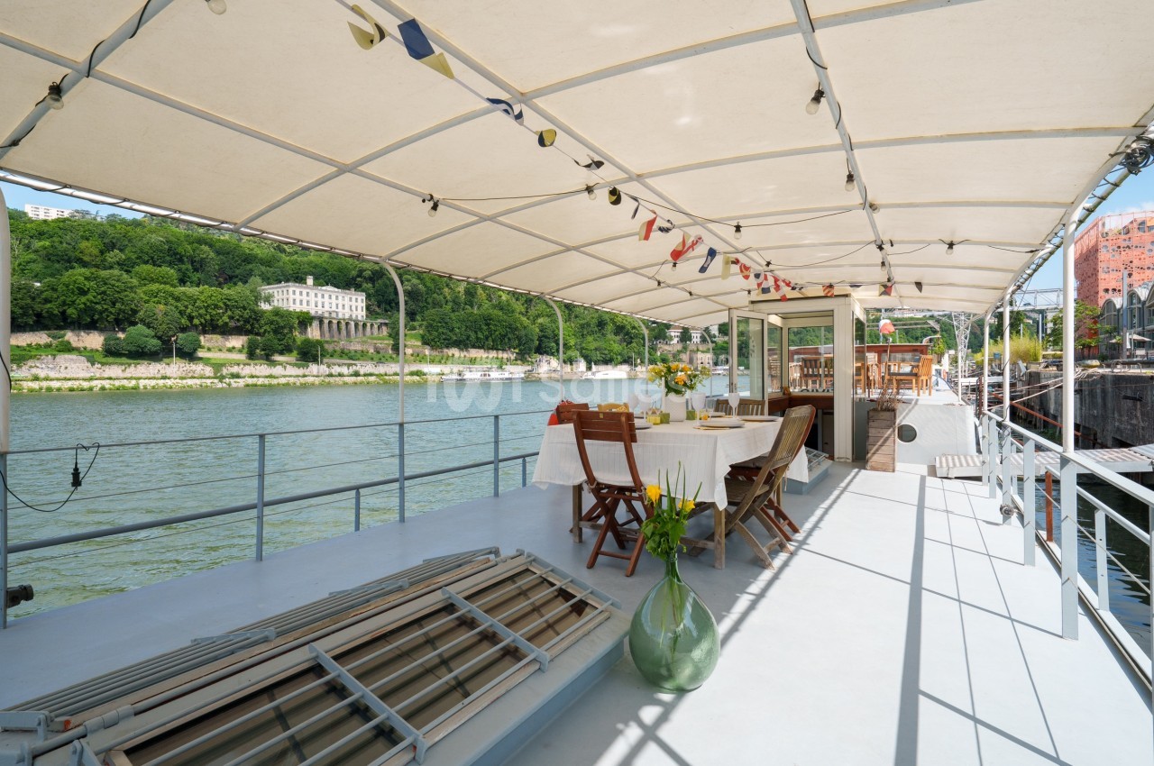 Terrasse couverte d'un bateau aménagée avec des tables dressées, chaises en bois et vue sur une rivière bordée de verdure.