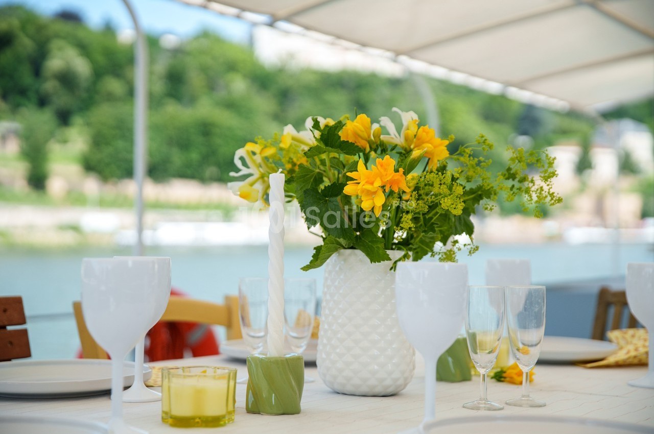 Table dressée en extérieur avec un vase de fleurs jaunes, des verres et des bougies, vue sur un paysage verdoyant.