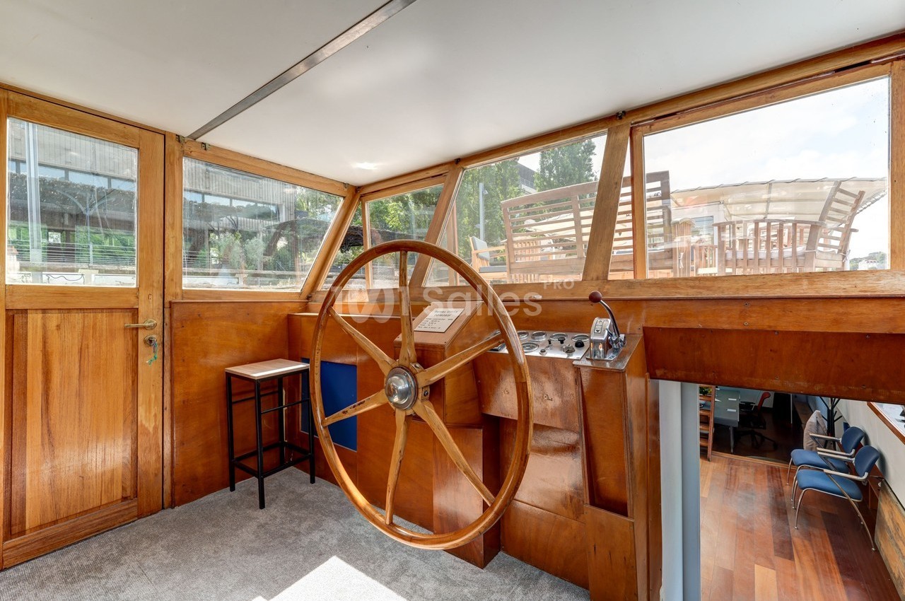 Poste de pilotage en bois d'un bateau avec un grand gouvernail, entouré de fenêtres offrant une vue extérieure.